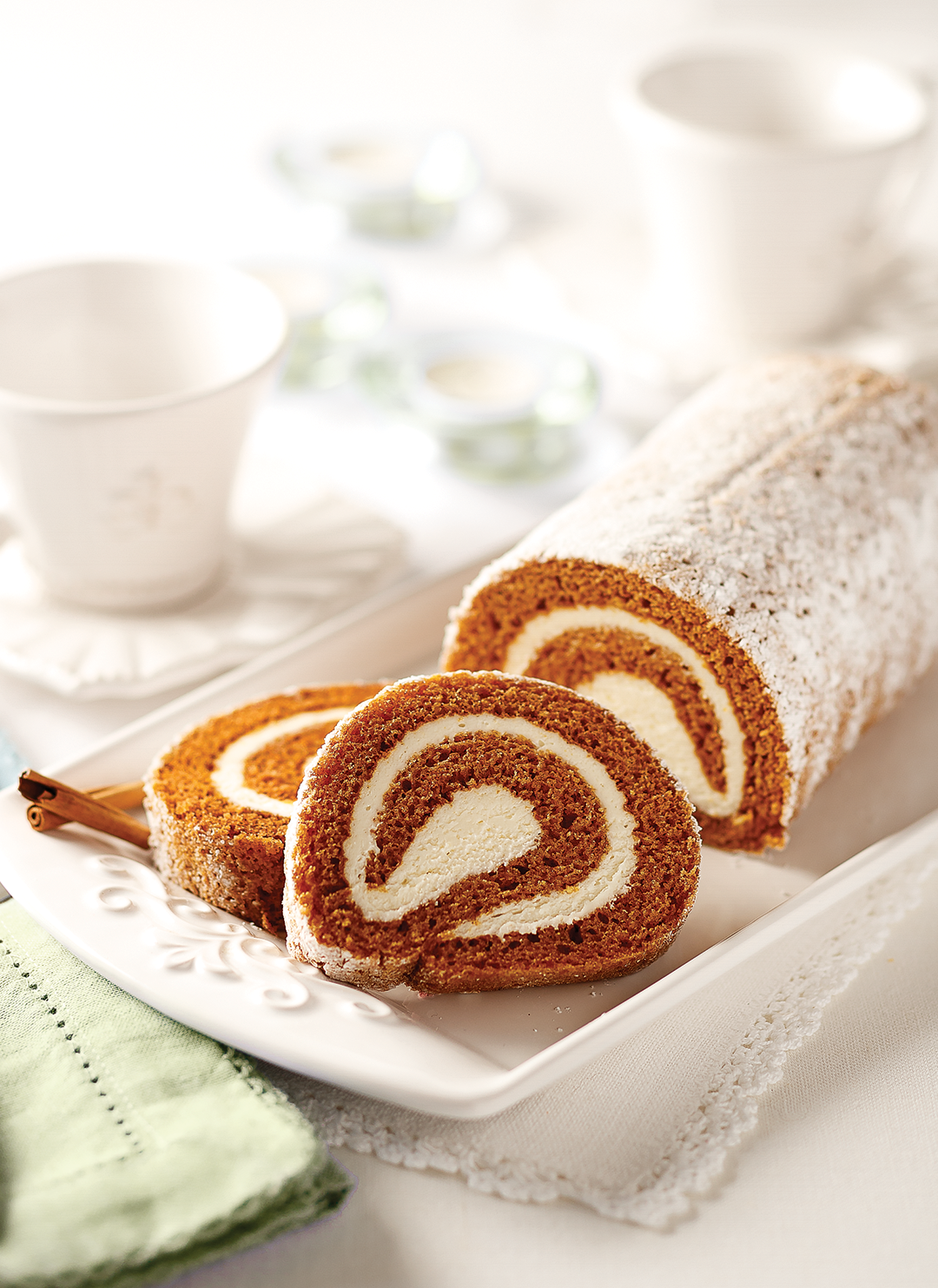 A sliced pumpkin spice cake roll with a cream swirl, dusted with powdered sugar and served on a white rectangular plate with a cinnamon stick, set on a white tablecloth with soft-focus white teacups in the background.