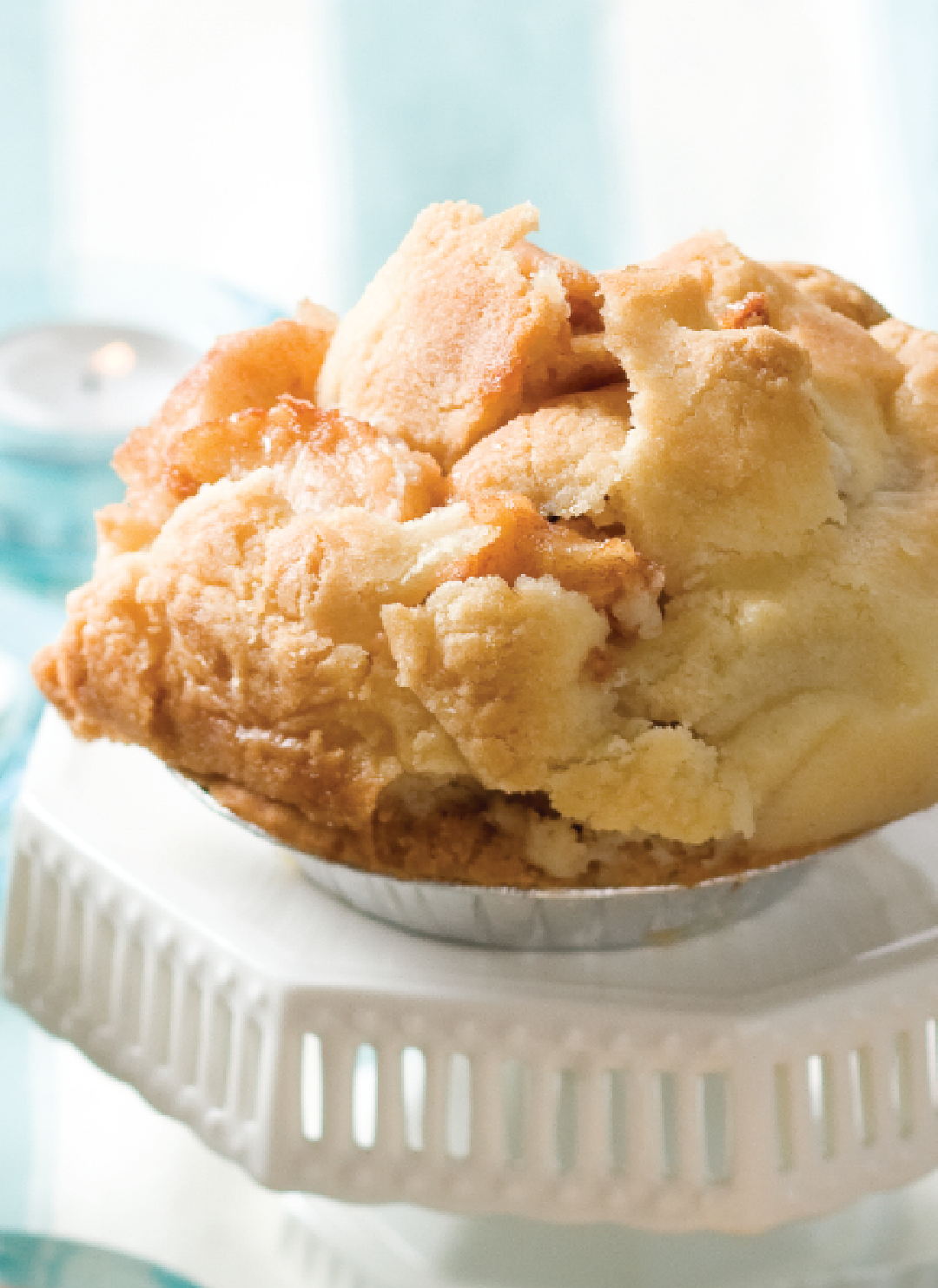A flaky crusted mini apple pie on a while plate with a soft blue background.